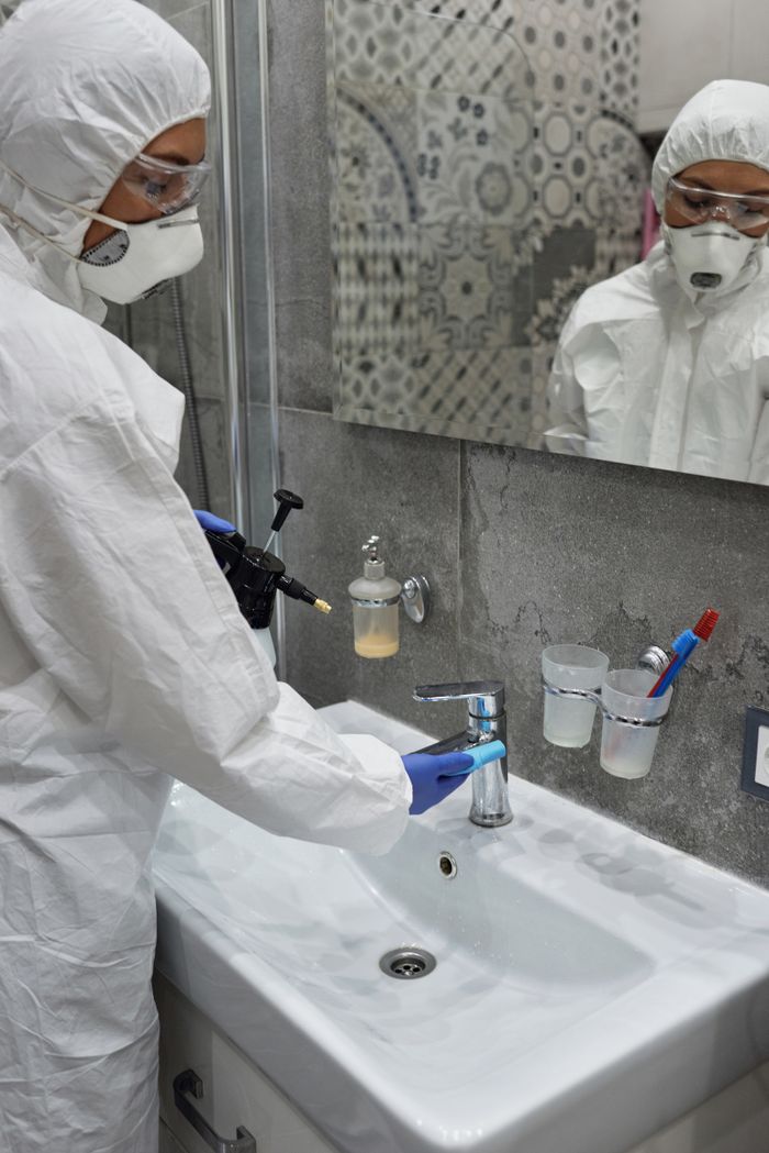 services-01 Person in protective gear disinfecting bathroom sink with spray bottle.