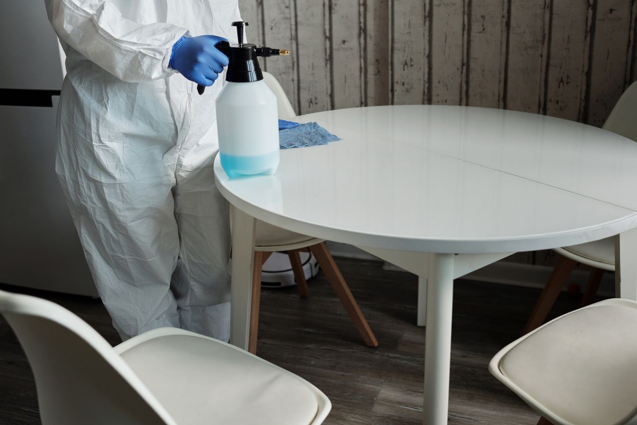 services-03 Person in protective suit disinfecting a white table indoors with a sprayer.