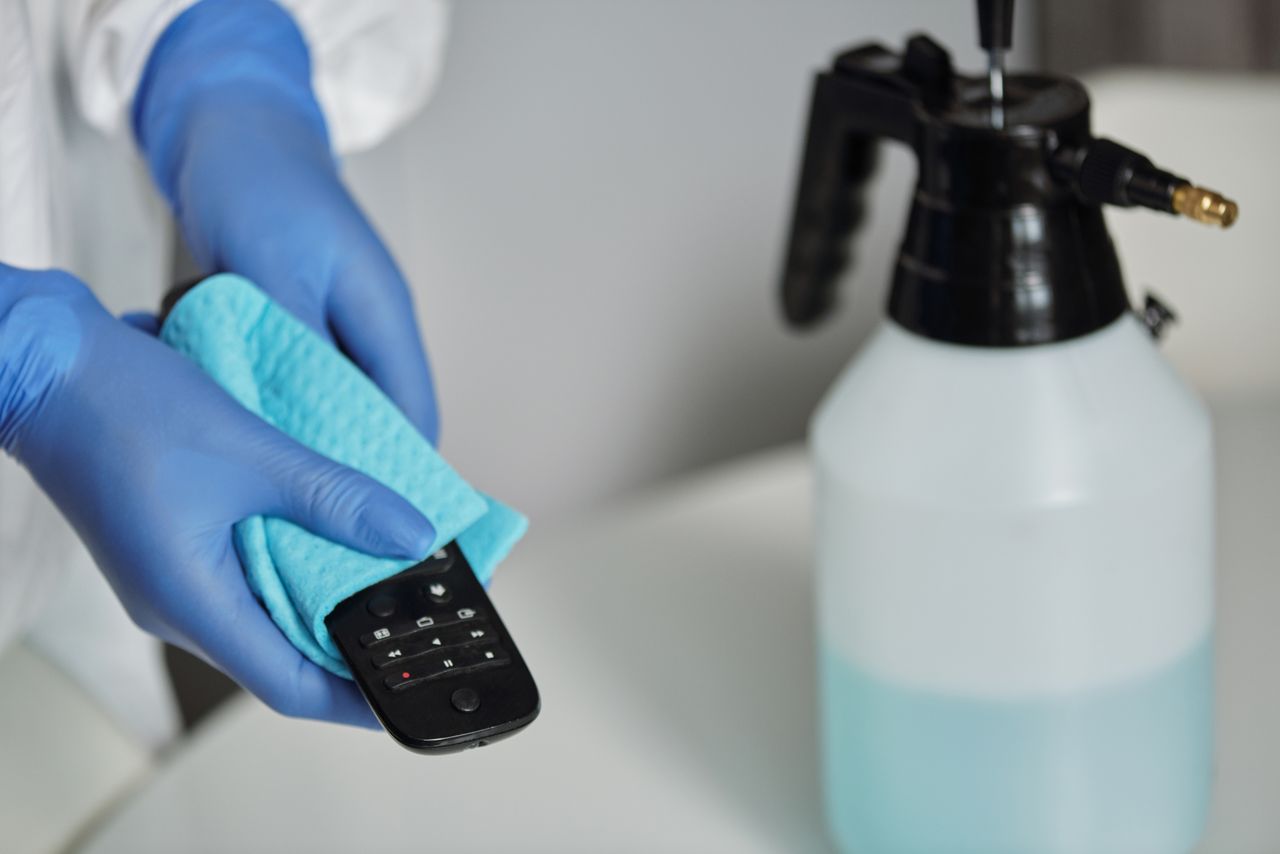 heros-img Close-up of gloved hands disinfecting a remote control with a cloth and spray bottle.